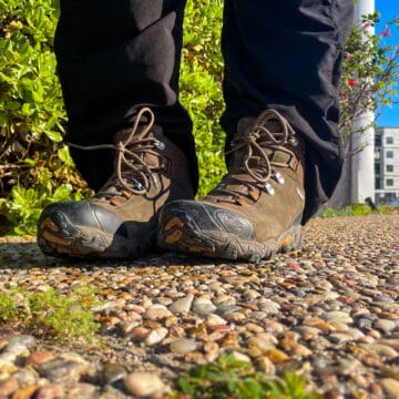 woman wearing oboz hiking boots as work boots near a construction site with bush in background, standing on pebble sidewalk