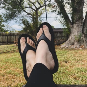 a woman wearing black pants and black oofos waterproof flip flops resting her legs on the side of a bench in a backyard